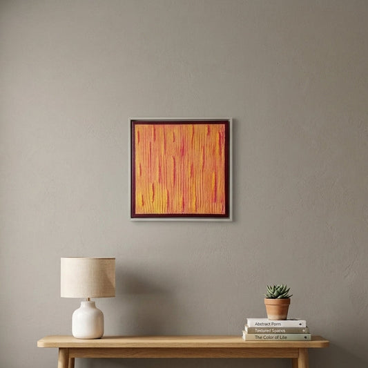 A framed abstract painting with vertical orange, yellow, and red brushstrokes hangs on a gray wall above a light wood console table in a styled room with a lamp, books, a small plant, and a woven basket.
